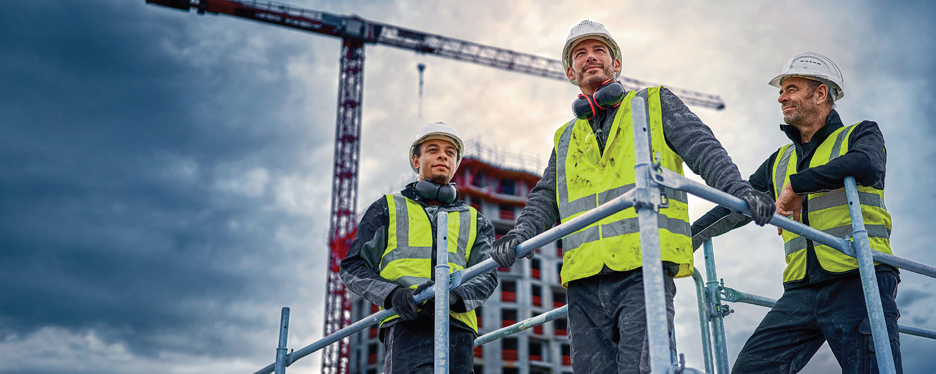 三个穿着安全背心的建筑工人在脚手架上，俯瞰起重机和建筑工地。