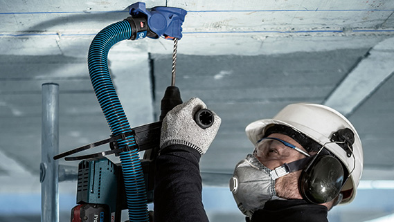 一名戴着头盔和耳罩的工人在天花板上钻孔。